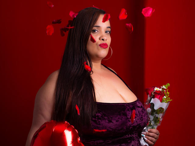AloondraZaenss with big tits in a purple velvet dress, holding flowers, red rose petals on face, long brown hair, red background, and shiny red balloon in left hand.