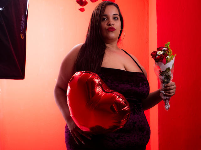 AloondraZaenss with large tits in a purple velvet dress, holding a red heart balloon and bouquet, red lipstick, long brown hair, standing against a red wall.