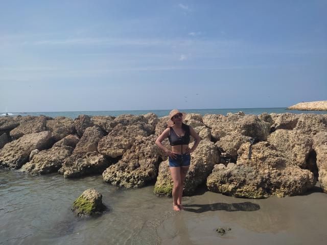 LunaCandy debout dans une eau peu profonde, vêtue d'un soutien-gorge de sport noir et de shorts bleus, les mains sur les hanches, pieds nus, plage rocheuse, ciel clair, légère peau bronzée, lunettes de soleil, décontractée, confiante..