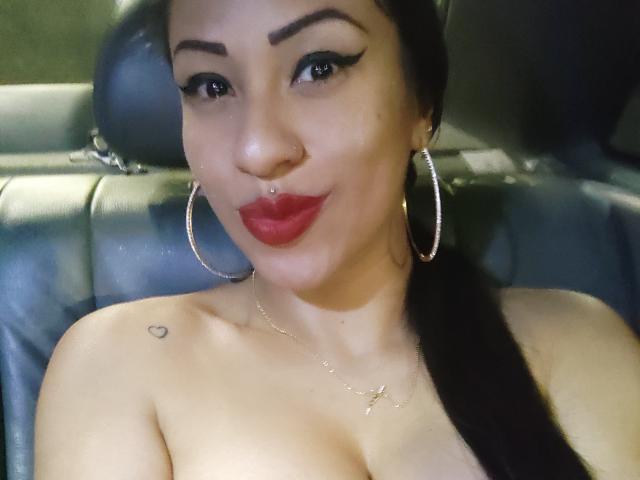 LunaCandy with dark hair, red lipstick, hoop earrings, nose piercing, small heart tattoo, necklace, and visible cleavage, sitting in a car. Her tits are slightly exposed.