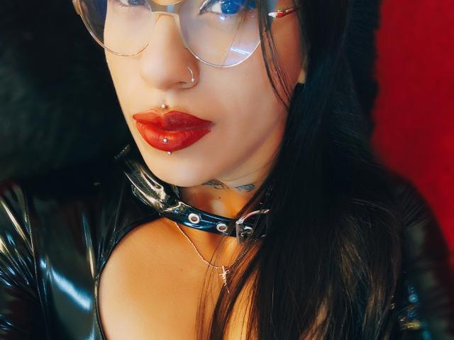 LunaCandy, con rossetto rosso, naso con piercing e occhiali, indossa una collana di pelle nera e una giacca lucida, ha un tatuaggio sul collo e un piercing al labbro..