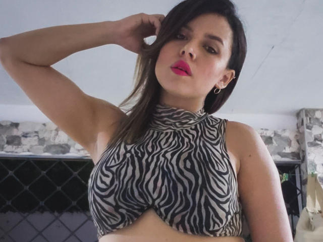 LindaRousse mit mittelgroßer Brust, in einem Crop-Top mit ZEBRA-Muster, mit pinkem Lippenstift, braunen Haaren und Reifenohrringen, die ihren Arm hebt und in die Kamera schaut..