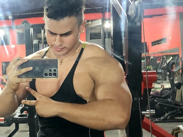 Muscular AngelLin with tan skin, black tank top, short dark hair, taking a mirror selfie in a gym, showing off bulging biceps and a gold cross necklace.