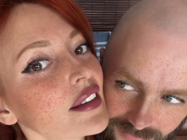 MaryAndJacky with red hair and purple lipstick, freckled face, next to bald guy with beard, green eyes, both looking at camera, close-up shot, his face partially in her.