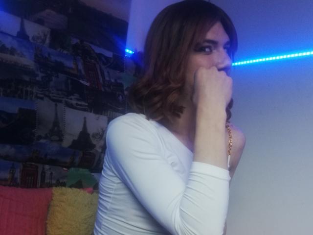 LunaGransTs with brown, wavy hair, white long-sleeve shirt, covering her mouth with her hand, blue LED strip light in the background, and a gold necklace.