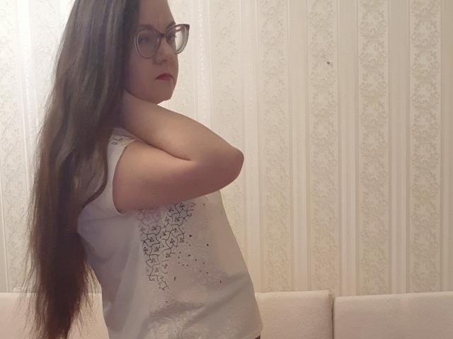 Anna Fock mit langen, braunen Haaren, Brille und rotem Lippenstift, trägt ein weißes Top ohne Ärmel und steht mit den Armen hinter dem Kopf, wobei kleine Brüste und eine dezente Hüftlinie zu sehen sind..