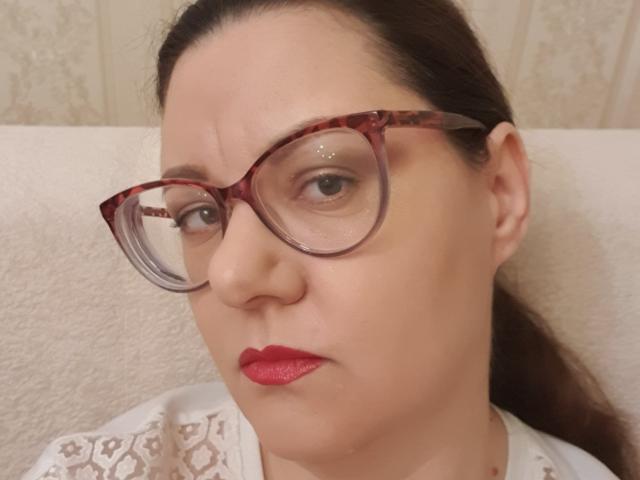 AnnaFock with tortoiseshell glasses, red lipstick, and a serious expression, wearing a white lace top, has a slightly wrinkled forehead and brown hair pulled back.