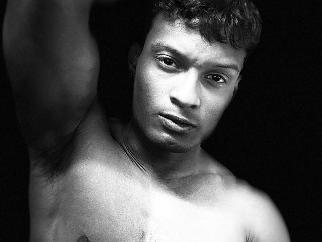 Foto em preto e branco de JacksonCooper com o braço levantado, mostrando pelos da axila, sem camisa, olhando ligeiramente para baixo com uma expressão séria, cabelo curto e encaracolado e pele lisa..