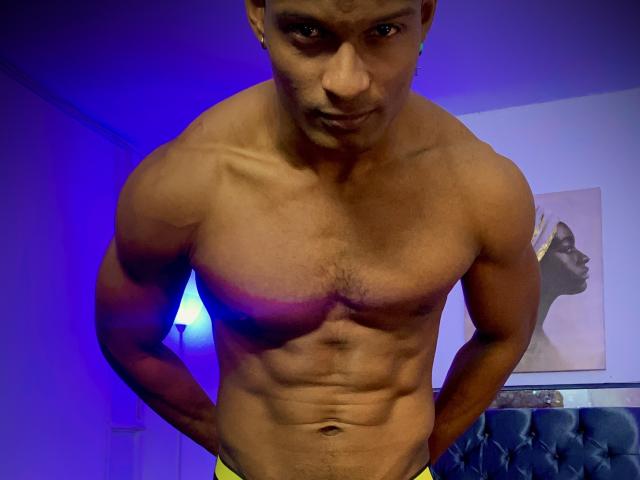 Shirtless JacksonCooper with a muscular, toned body, visible abs, and hands on yellow pants, looking down with a slight smirk. Blue light in the background.