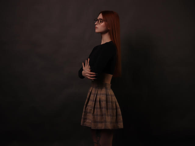 MalenaGolden, avec de longs cheveux roux, des lunettes, un haut noir et une jupe à carreaux, debout de côté, les bras croisés, montrant de petites seins, fond sombre, regardant vers le haut, éclairage sombre..