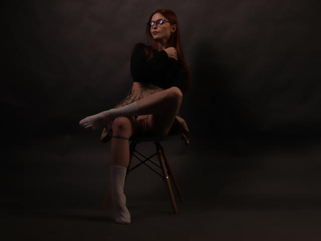 Malena, met een bril, bruin haar, een zwarte top, een beige rok, witte sokken en gekruiste benen, zittend op een stoel, donkere achtergrond, rechterknie omhoog, linkerknie naar beneden, naar boven kijkend..