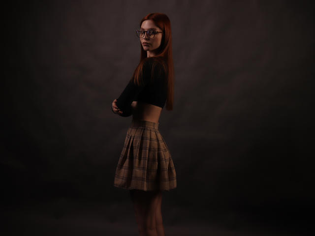 Malena, met rood haar, een bril, een zwart topje en een geruite rok, staat zijwaarts, met armen over elkaar, op een donkere achtergrond. Haar achterkant is zichtbaar, ze heeft lange benen en een serieuze uitdrukking..