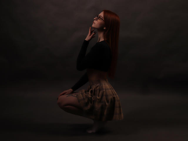 Malena, cheveux roux, lunettes, crop top noir, jupe à carreaux, accroupie, main près du visage, fond sombre, regardant vers le haut, manches longues, mignonne, sexy, pieds nus, éclairage doux..