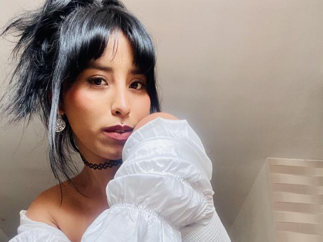 ElizabethMacy with messy black hair, white off-shoulder shirt, choker, nose ring, and earrings, resting her head on her hand, looking at the camera with a slight pout.