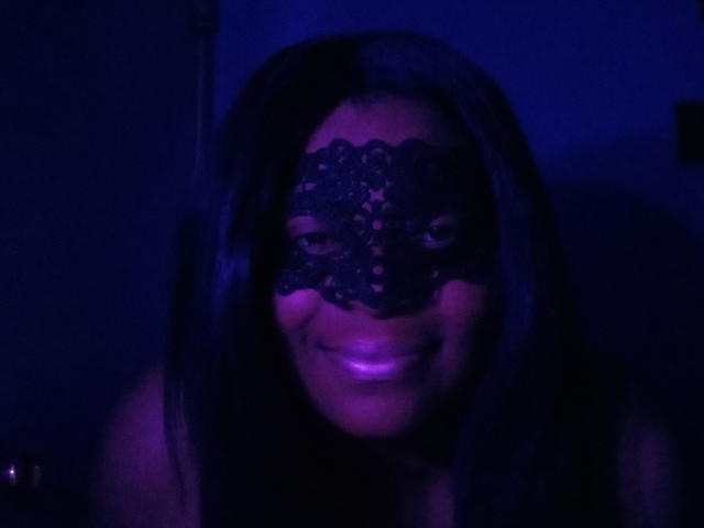 BettyBop with dark hair, wearing a black lace mask, smiling in blue-purple light, her shiny tits slightly visible in the dim light. Her eyes are visible through the mask.