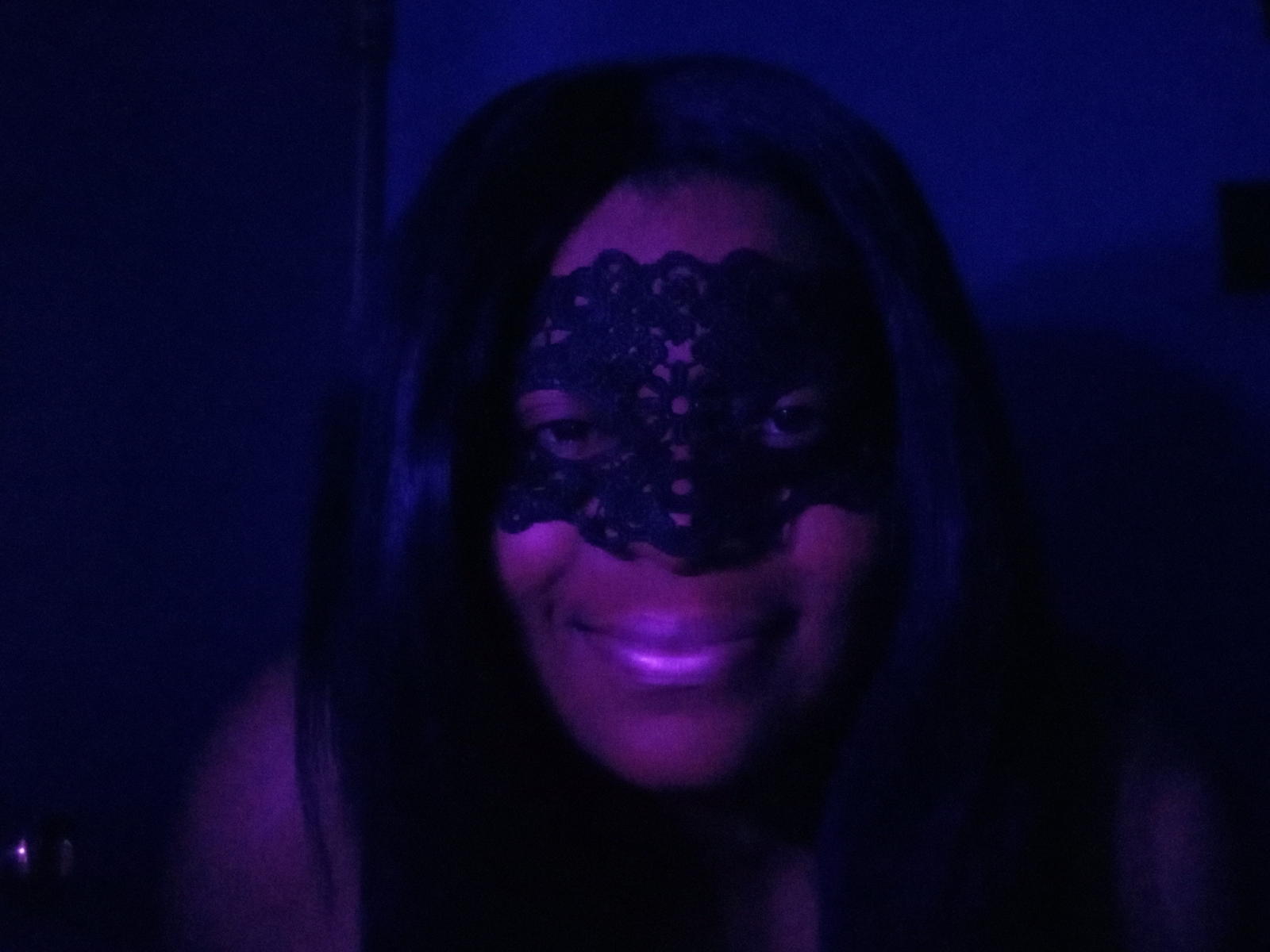 BettyBop with dark hair, wearing a black lace mask, smiling in blue-purple light, her shiny tits slightly visible in the dim light. Her eyes are visible through the mask.