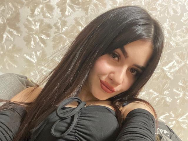 SofiaCrazy with long black hair, red lipstick, and black off-shoulder top, smiling softly at the camera, showing her tits slightly. She's leaning on a gray chair.