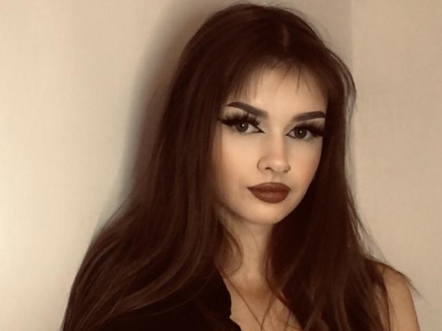 VickyxFantasy, con cabello largo y castaño, lápiz labial marrón oscuro y pestañas dramáticas, vestida con una camisa negra, mirando ligeramente hacia un lado con una sonrisa sutil..