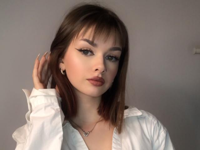 VickyxFantasy has a cute face with sharp eyeliner, brown bob, white shirt, and a necklace with a small heart. She's touching her hair with a serious expression.