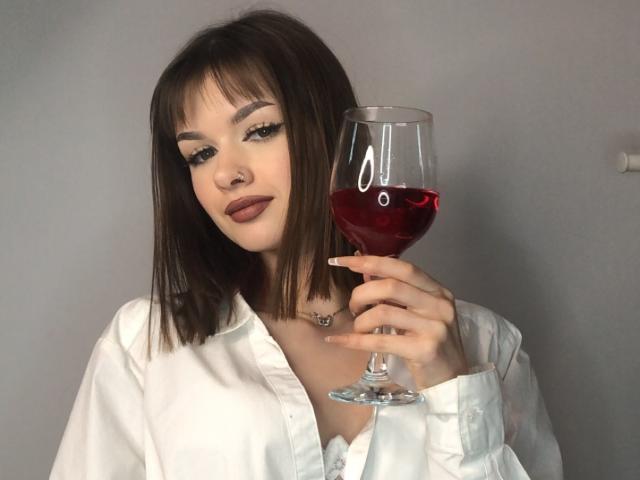 VickyxFantasy con un taglio di capelli corto marrone liscio, indossa una camicia bianca, tiene un bicchiere di vino rosso, con un sorriso sornione, ha un piercing al naso, un trucco leggero, sfondo grigio. Il seno è parzialmente visibile, di piccola o media taglia..