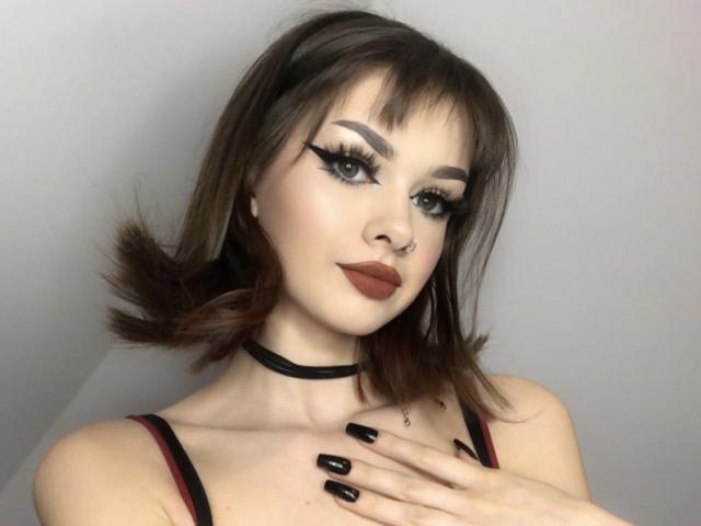 VickyxFantasy, con i capelli castano scuro, un collare nero, eyeliner ad ali di farfalla, rossetto scuro, unghie nere e un piercing al naso, che si tocca il petto..