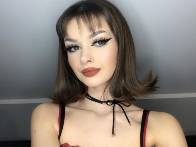 VickyxFantasy, com cabelo castanho na altura dos ombros, sobrancelhas grossas, delineador preto, batom vermelho, colar e um sutiã preto e vermelho, olhando para a câmera com um leve sorriso. Seios visíveis..