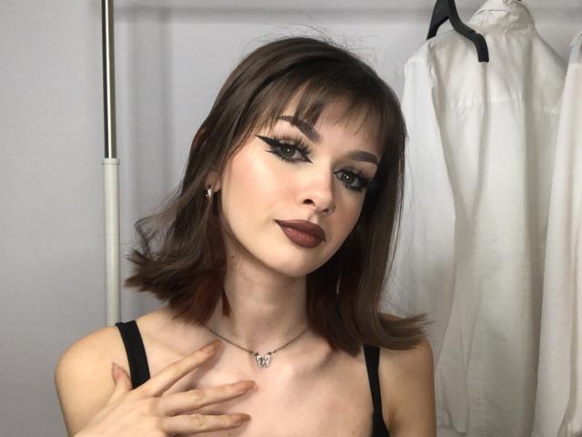 VickyxFantasy tiene el cabello castaño hasta los hombros, delineador de ojos negro, lápiz labial oscuro, un piercing en la nariz y un collar con un pequeño dije, tocándose el pecho con una mano..