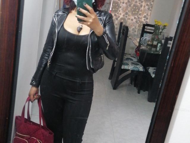 VioletaSwitch com cabelo ruivo, jaqueta de couro preta, blusa preta, calça preta, segurando uma bolsa vermelha, selfie no espelho, mostrando os seios, segurando um celular, esmalte preto, quarto com móveis..