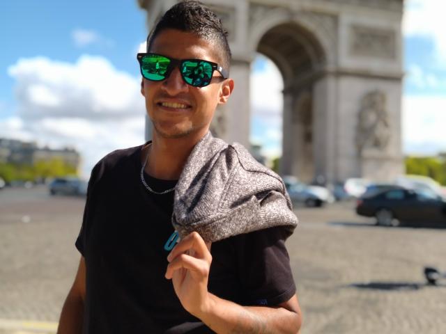 Vergara, con capelli corti e scuri, occhiali da sole verdi riflettenti, maglietta nera, sciarpa grigia di lana, sorridente, con una mano sulla spalla, indossa una collana a catena, in piedi in una zona soleggiata con strade acciottolate, un arco sullo sfondo..