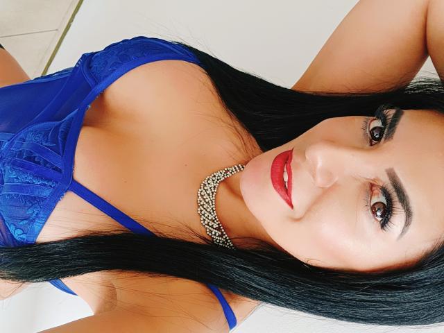 JoTaylor with long black hair, blue lace bra, red lipstick, and a diamond necklace, showing her tits in a selfie, smiling with red lips and dark eyebrows.