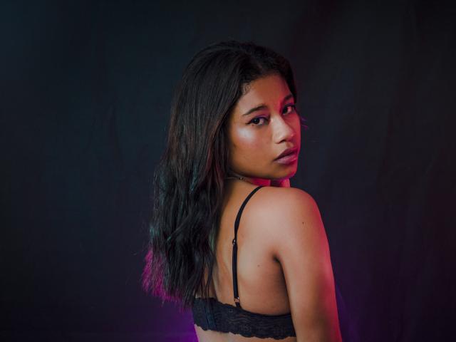 TamiDubois, met donker, golvend haar, draagt een zwarte kanten bh, kijkt over haar schouder, heeft een glanzende huid, een medium bruine huidskleur en een subtiel roze en paarse gloed op haar rug..