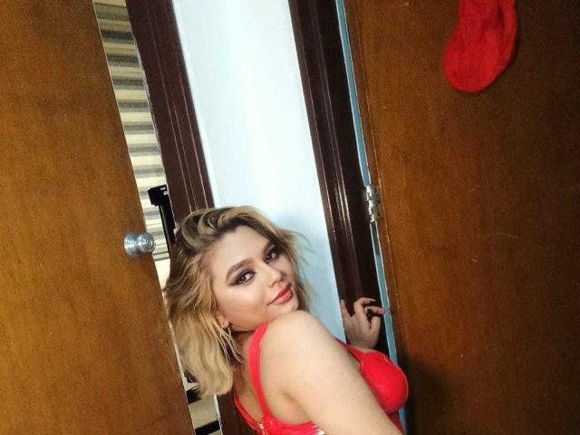 AliceeSecret, com cabelo loiro, sutiã vermelho e batom vermelho, olhando por cima do ombro, em pé em uma porta, mostrando os seios, com uma meia vermelha na porta..
