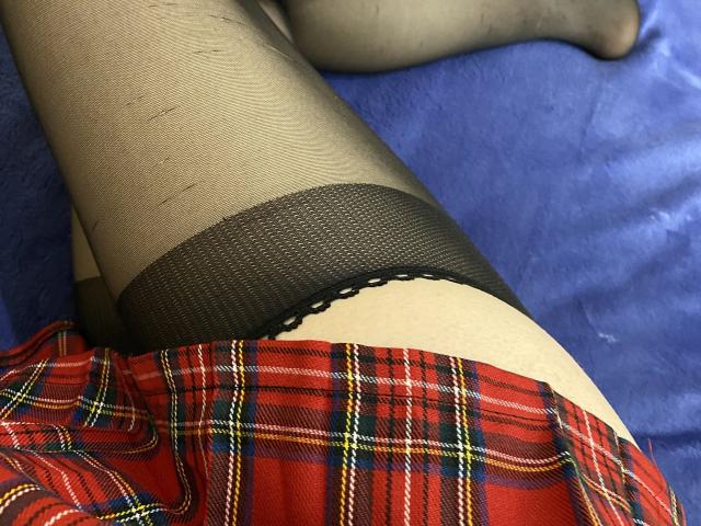 Rubbi's black mesh stockings with lace top, red plaid skirt, and pale skin are visible. Her legs are crossed on a blue surface. Her pussy is not shown.