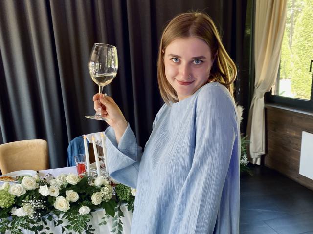 SexyBreakss with light brown hair, blue eyes, and a cute smile, holding a wine glass, wearing a light blue, textured shirt, sitting at a table with flowers.