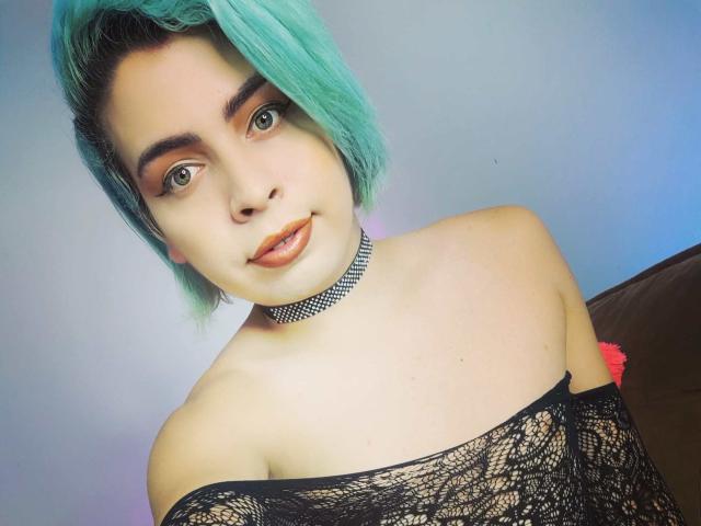TsFoxCandy with teal hair, wearing a black lace off-shoulder top, choker, and orange lipstick, showing off her tits. She's looking at the camera with a subtle smile.