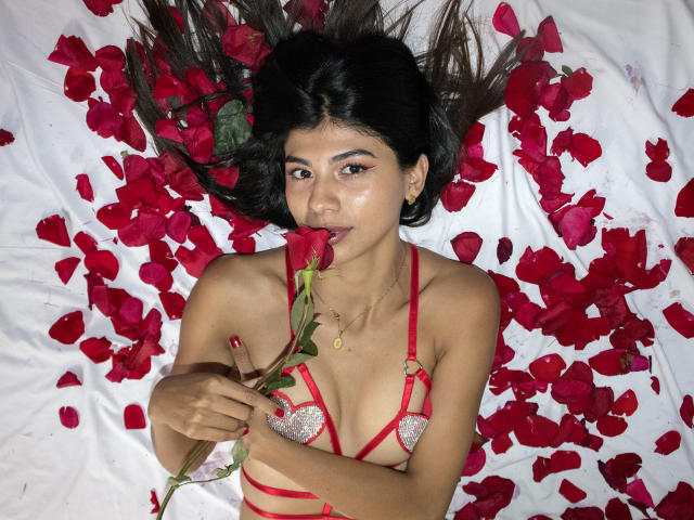 SamyNicols with dark hair, wearing red strappy lingerie, lying on white with red rose petals, holding a red rose to her lips, looking at the camera.