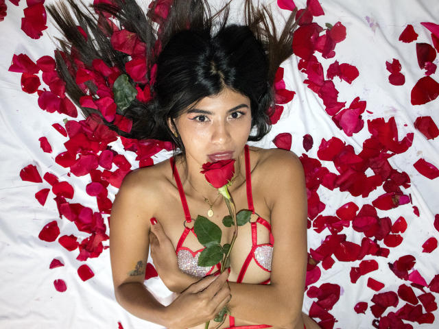 SamyNicols with dark hair, wearing red lace bra, lying on white bed with red rose petals, holding red rose, looking at camera, tits visible, tattoo on right arm.