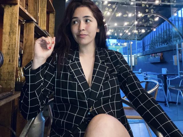 Nico Coleman, con cabello oscuro y ondulado, vistiendo un blazer a cuadros negros y blancos, sosteniendo un cigarrillo, sentado en una silla, mirando hacia un lado con una ligera sonrisa..