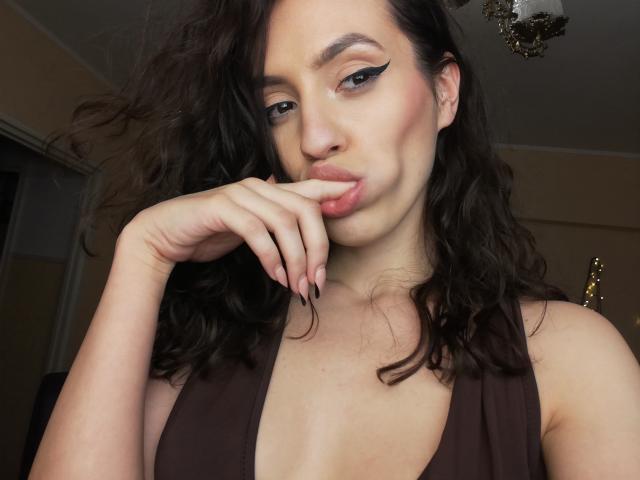 VievienneClemente with wavy dark brown hair, winged eyeliner, and pink lipstick, biting her finger, wearing a brown deep-pleated top, showing her small tits.