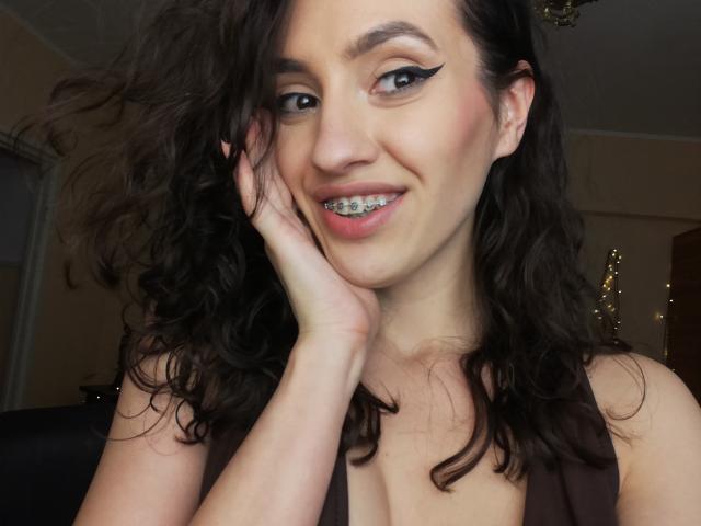VievienneClemente has curly black hair, bold eyeliner, and a tooth grill, smiling with her hand on her cheek, showing off her tits in a brown top.