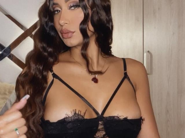 ValeriaKlaus with long wavy brown hair, wearing a black lace bralette, showing her big tits, and a red pendant necklace, pouting with full lips.