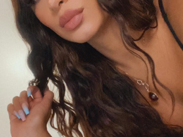 ValeriaKlaus with pouty lips, long wavy brown hair, blue nails, holding hair, wearing a necklace with a brown pendant, black strap visible, soft light, close-up.