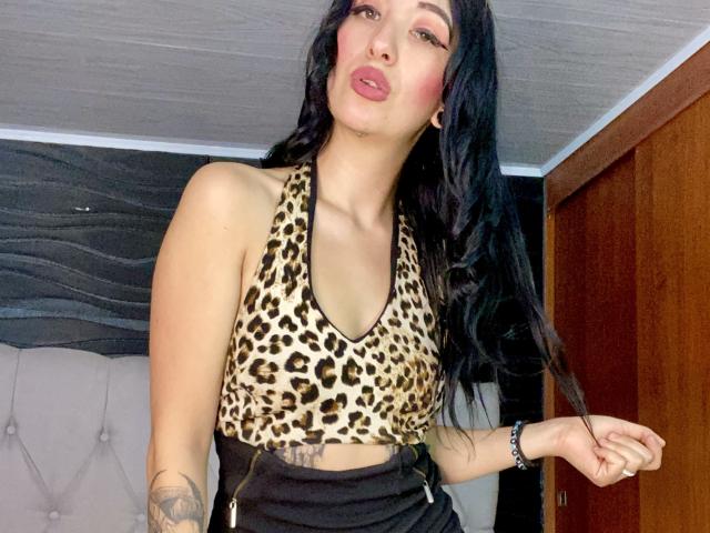 TifanyDiaz with long black hair, pouty pink lips, wearing a leopard print top, showing her small tits, and black high-waisted pants, has tattoos on her right arm.