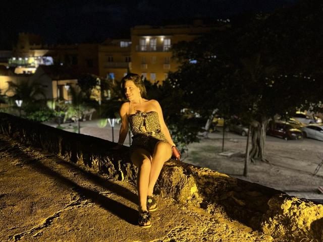 Liliysweet sitting on a rough stone ledge at night, wearing a strapless leopard print top and black shorts, with her legs crossed and a serious expression on her face.