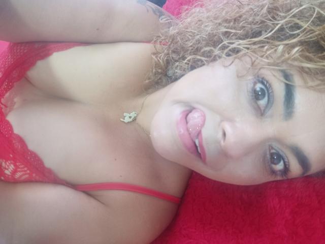 SolmaryHot with curly blonde hair, red lace top, and tongue out, looking at camera, showing tits, pink lipstick, and a gold necklace, lying on a red textured surface.