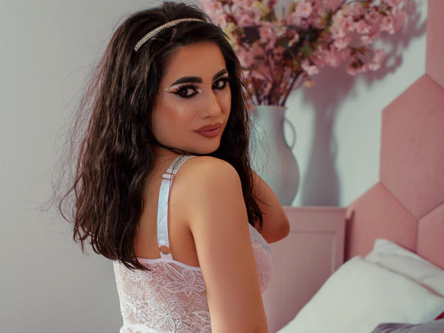 EmaBrownie with wavy dark hair, wearing white lace lingerie, looking back with a slight smile, showing her tits, in a pink bedroom with a vase of pink flowers.