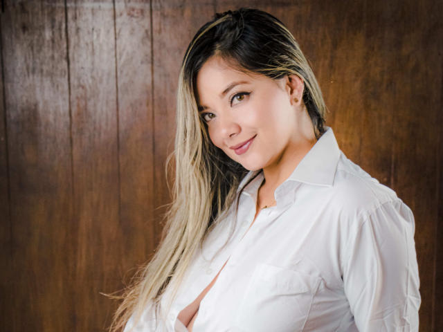 AbrilClark, con cabello largo rubio y negro, vestida con una camisa blanca desabrochada que revela sus pequeños senos, sonriendo a la cámara, de pie frente a una pared de madera..