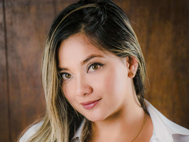 AbrilClark's face, featuring brown eyes, light skin, and blonde-streaked black hair, with a subtle smile, wearing a white shirt, small ear piercing visible.