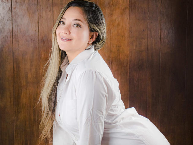 AbrilClark with long blonde hair, white shirt, smiling, looking over shoulder, brown wood background, cute, white earrings, subtle makeup, white shirt buttons, slight blush, relaxed, confident.