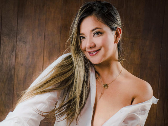 AbrilClark met lang blond haar, lachend, gekleed in een wit shirt dat van haar schouder valt, waarbij haar borsten zichtbaar zijn, draagt een gouden ketting, op de achtergrond een houten oppervlak..